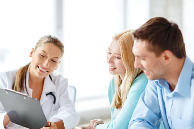 Picture of a female Physician holding up a clipboard, pointing at it, and smiling while she is talking to a couple that is also smiling as they look down at the clipboard.