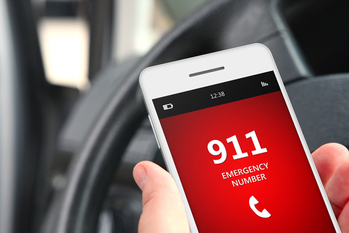 Picture of a person&apos;s hand holding a smart phone out in front of a car steering wheel. The phone&apos;s screen says: 911 EMERGENCY NUMBER