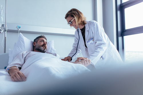 Picture of a female Physician leaned over checking a male patients heartbeat. The male patient is lying down in a swing bed in a hospital room.